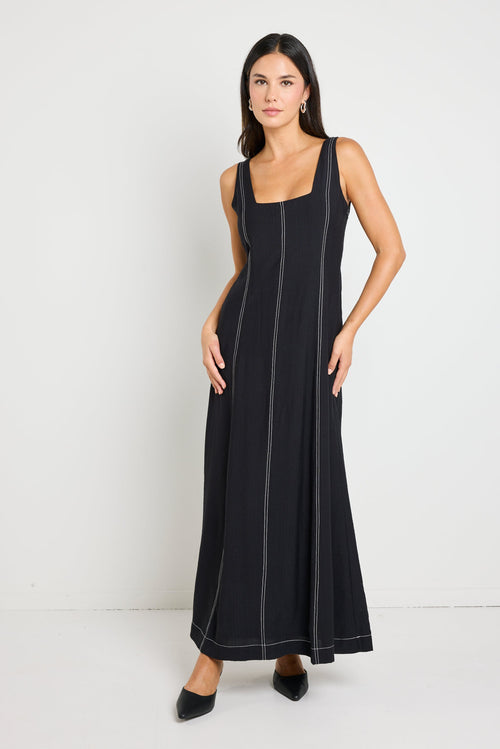 Woman wearing a black maxi dress with a square neckline and contrast stitching, styled with black heels for a chic, modern look.