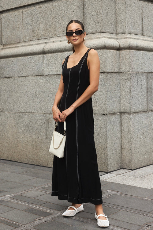 Woman wearing a black maxi dress with a square neckline and contrast stitching, styled with black heels for a chic, modern look.