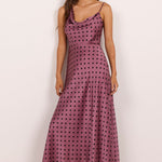 A woman modeling a sleeveless, floor-length cowl-neck dress with a dusty rose base and black polka dots, paired with black flat sandals, set against a plain off-white studio background.