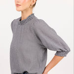 A woman modeling a gray and white micro-check patterned blouse with a ruffled mock neck and elbow-length puff sleeves, paired with blue jeans against a plain white studio background.