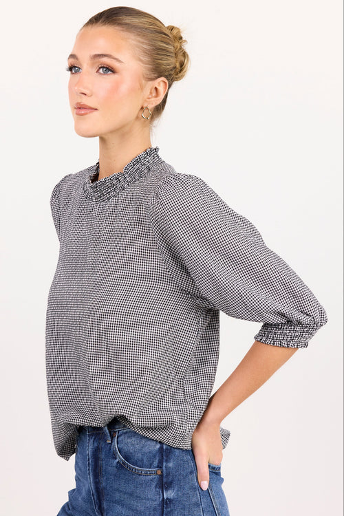 A woman modeling a gray and white micro-check patterned blouse with a ruffled mock neck and elbow-length puff sleeves, paired with blue jeans against a plain white studio background.