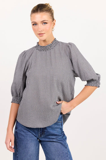 A woman modeling a gray and white micro-checkered blouse with a textured embroidered yoke and rolled three-quarter sleeves, paired with blue jeans against a plain white studio background.