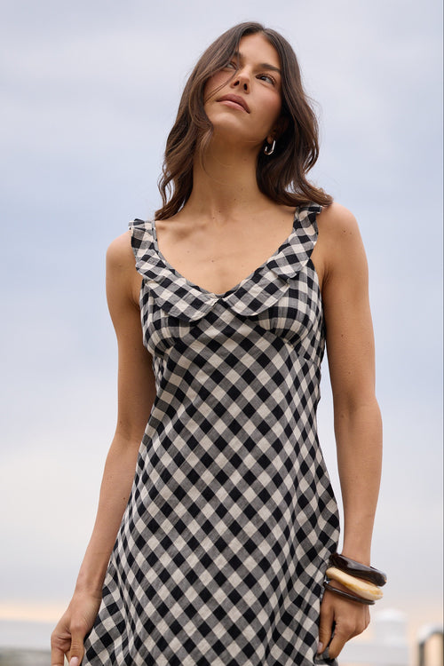 A woman wearing a black and white gingham check sleeveless dress, photographed outdoors against a soft-focus sky and distant water background.
