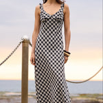 A woman stands on a concrete pier by the ocean wearing a black and white checkered sleeveless maxi dress. The background features a calm sea, rocky shoreline, and a cloudy sky with a soft sunset glow.