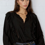 Woman wearing a black blouse and blue jeans against a white brick wall.