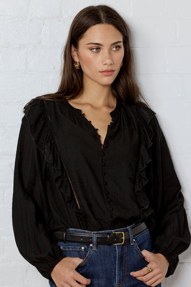 Woman wearing a black blouse and blue jeans against a white brick wall.