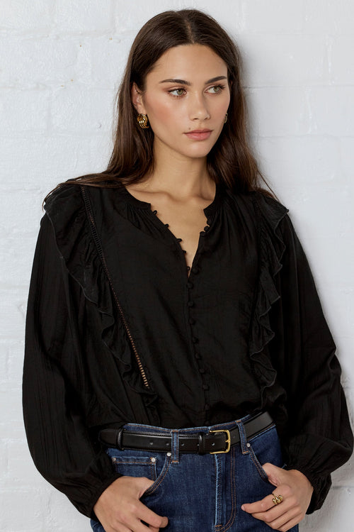 Woman wearing a black blouse and blue jeans against a white brick wall.