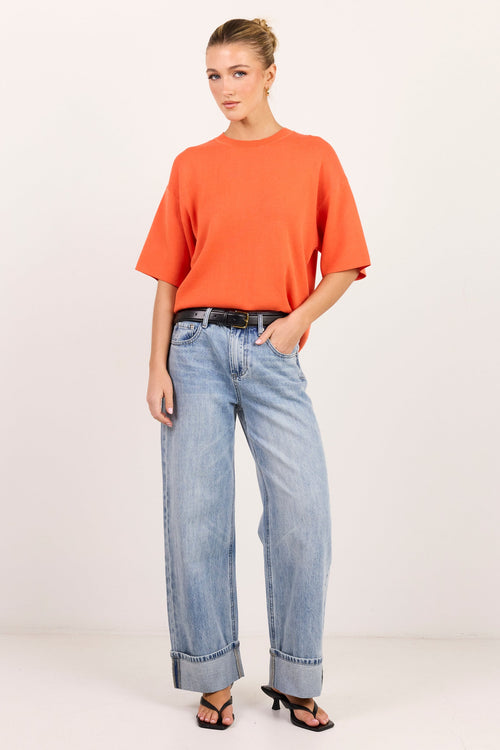 A woman modeling a bright orange short-sleeve crewneck top, light-wash wide-leg blue jeans, and black sandals against a plain white studio background.