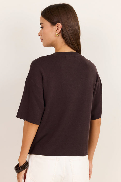 A woman shown from the back wearing a dark brown short-sleeve knit top and white pants against a plain off-white studio background.