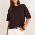 A woman modeling a dark brown short-sleeve knit top with white pants, standing against a plain off-white studio background.