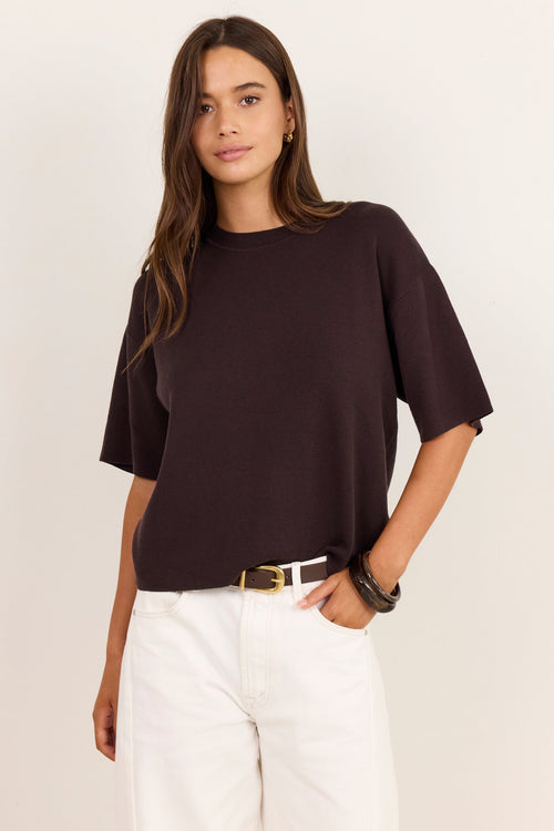 A woman modeling a dark brown short-sleeve knit top with white pants, standing against a plain off-white studio background.