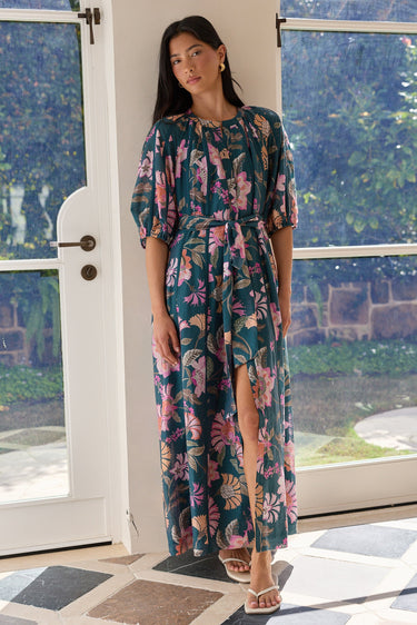 A woman modeling a dark teal maxi dress with a pink and green tropical floral print, standing in a bright doorway with a garden view.