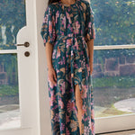 A woman models a floral midi dress with a high slit, standing in a doorway with a garden view in the background. The dress features a dark teal base with pink, purple, and green tropical floral patterns.