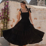 Woman wearing a black tiered maxi dress with shirred bodice and bow-tie straps, styled with statement earrings and woven black slides.