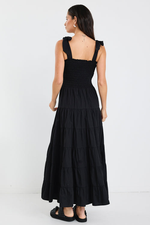Woman wearing a black tiered maxi dress with shirred bodice and bow-tie straps, styled with statement earrings and woven black slides.