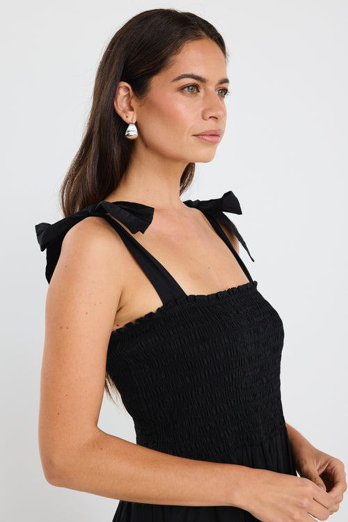 Woman wearing a black tiered maxi dress with shirred bodice and bow-tie straps, styled with statement earrings and woven black slides.