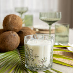 A white candle in a clear glass holder with a delicate green botanical leaf pattern, styled on a table with palm leaves, coconuts, and green glassware in the background.