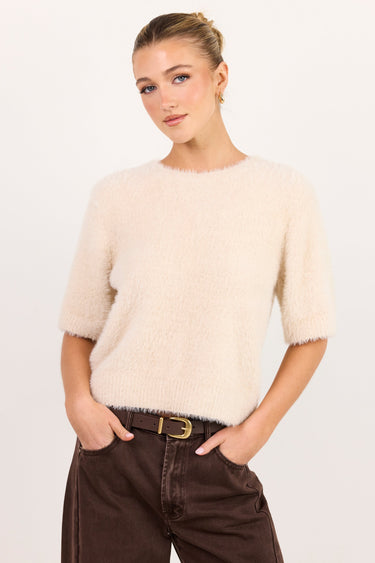 A woman modeling a short-sleeved, fuzzy cream-colored knit sweater paired with dark brown wide-leg jeans and a gold-buckled belt, set against a plain light gray studio background.
