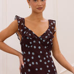 A woman wearing a dark brown sleeveless dress with a light blue polka dot pattern and ruffled shoulder details, posing in an indoor setting with white paneled walls.