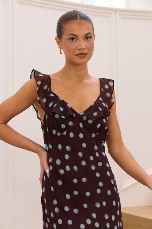 A woman wearing a dark brown sleeveless dress with a light blue polka dot pattern and ruffled shoulder details, posing in an indoor setting with white paneled walls.