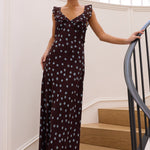 A woman wearing a sleeveless dark brown maxi dress with a light blue polka dot pattern stands on wooden stairs inside a bright white building, holding onto a black metal railing.