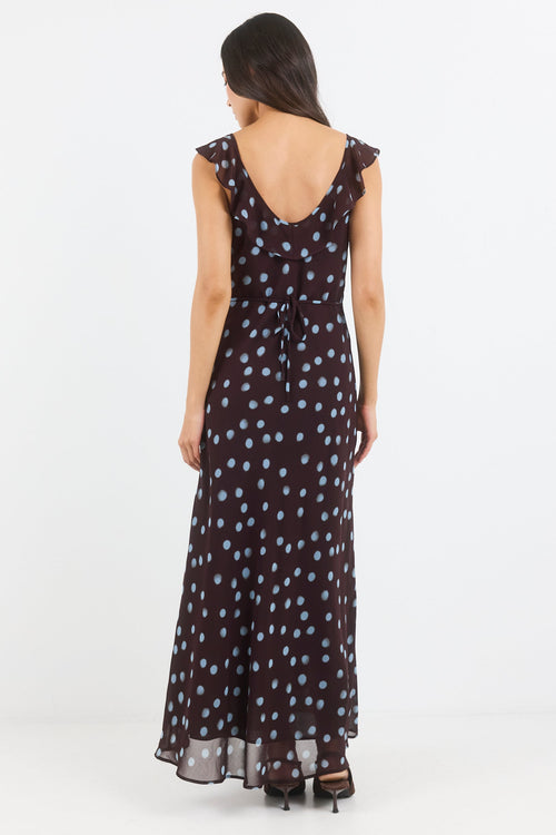 A woman viewed from the back wearing a sleeveless dark maxi dress with a small light polka dot pattern and a ruffled open back, photographed against a plain light gray studio background.