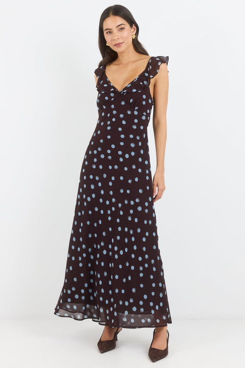 A woman modeling a sleeveless black maxi dress with a white and light blue polka dot pattern, ruffled shoulder straps, and a V-neckline, set against a plain white studio background.