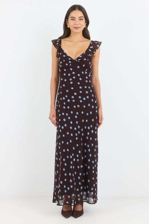 A woman modeling a sleeveless, V-neck maxi dress with a dark base and small white polka dots, standing against a plain white studio background.
