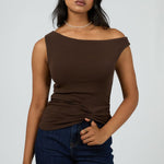 Woman wears a fitted, sleeveless chocolate brown top with an off-shoulder neckline, paired with dark denim jeans and minimal jewellery for a sleek, modern vibe.