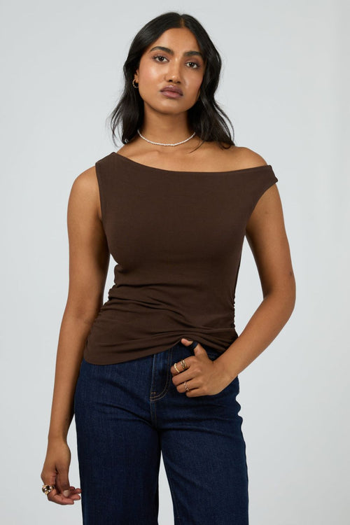 Woman wears a fitted, sleeveless chocolate brown top with an off-shoulder neckline, paired with dark denim jeans and minimal jewellery for a sleek, modern vibe.