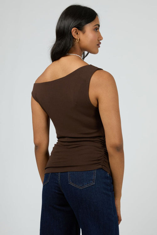 Woman wears a fitted, sleeveless chocolate brown top with an off-shoulder neckline, paired with dark denim jeans and minimal jewellery for a sleek, modern vibe.