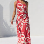 A woman models a red and white tropical leaf print maxi dress in an outdoor setting next to a swimming pool, with a white wall and green trees in the background.