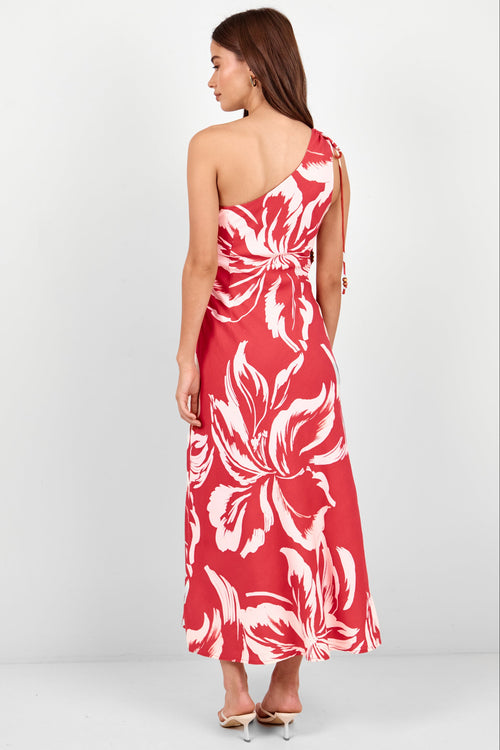 A woman modeling a red one-shoulder maxi dress with a large white tropical floral print, shown from a rear three-quarter angle against a light gray studio background.