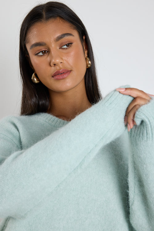 Woman wearing a soft sea foam green relaxed crew neck jumper styled with a silky white skirt, leaning against a concrete column surrounded by greenery.