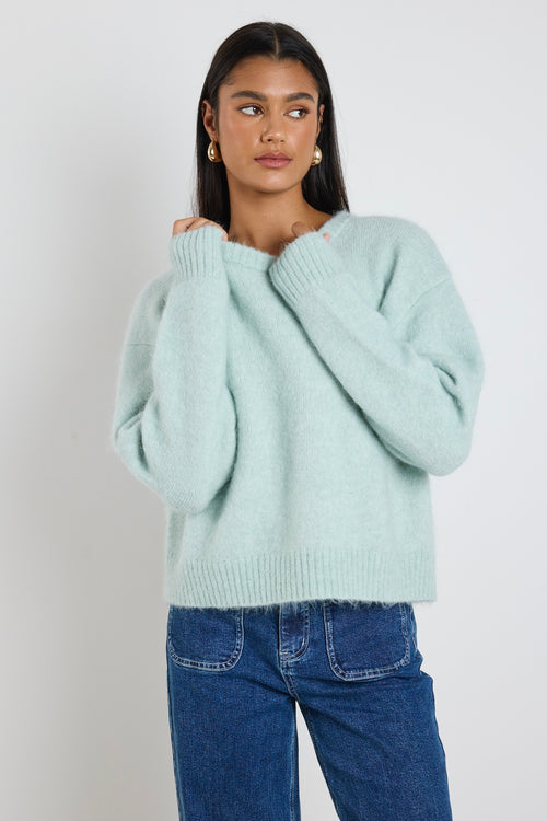 Woman wearing a soft sea foam green relaxed crew neck jumper styled with a silky white skirt, leaning against a concrete column surrounded by greenery.