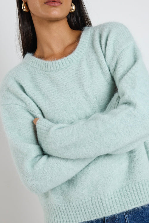 Woman wearing a soft sea foam green relaxed crew neck jumper styled with a silky white skirt, leaning against a concrete column surrounded by greenery.