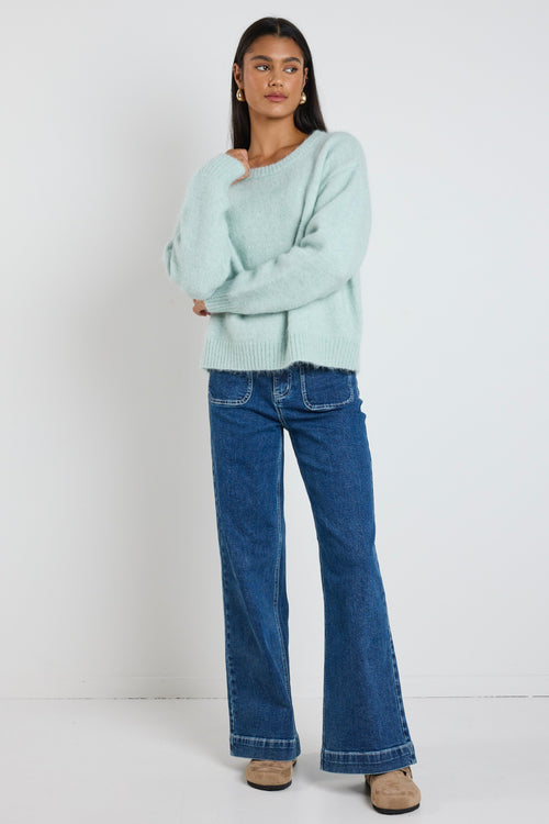 Woman wearing a soft sea foam green relaxed crew neck jumper styled with a silky white skirt, leaning against a concrete column surrounded by greenery.