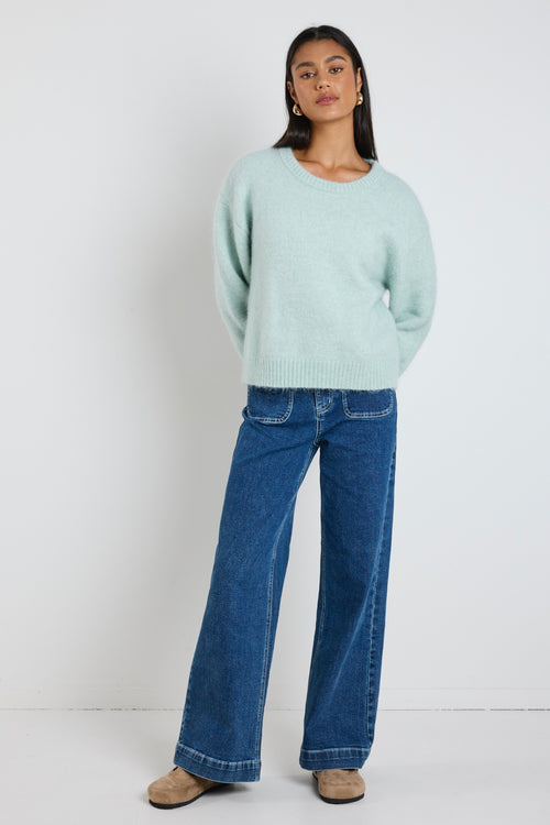 Woman wearing a soft sea foam green relaxed crew neck jumper styled with a silky white skirt, leaning against a concrete column surrounded by greenery.