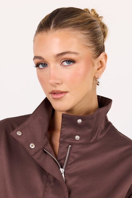 A woman wearing a brownish-rose high-collar zip-up jacket with silver snap buttons, shown from the chest up against a plain light background.