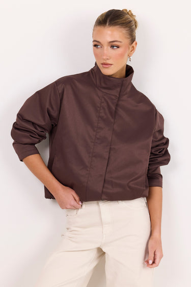 A woman modeling a cropped, high-neck jacket in a dark brown satin-like fabric, paired with cream-colored trousers, against a plain white studio background.