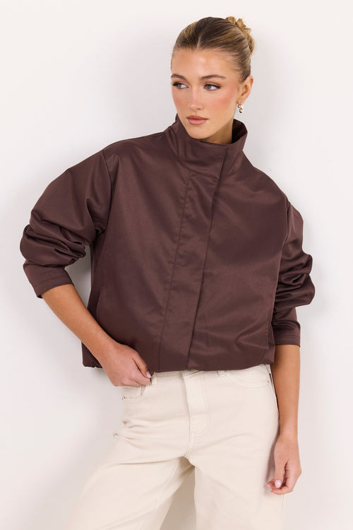 A woman modeling a cropped, high-neck jacket in a dark brown satin-like fabric, paired with cream-colored trousers, against a plain white studio background.