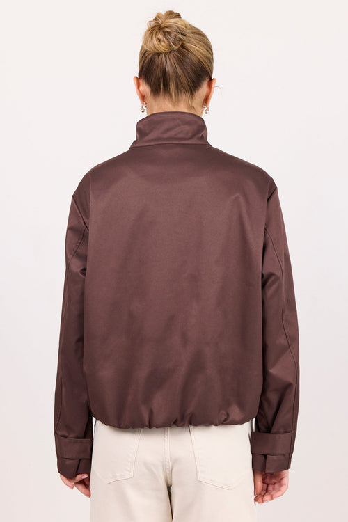 A woman viewed from behind wearing a dark brown satin-like jacket and cream-colored pants, standing against a plain white studio background.