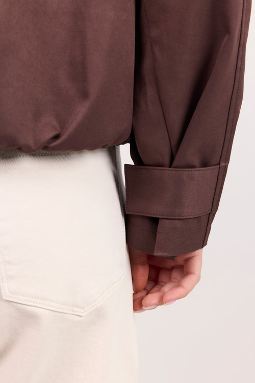 Close-up of a person wearing a dark brown cropped jacket with a rolled cuff and a cream-colored skirt, showing the fabric texture and fit against a light background.