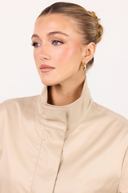 A woman with her hair in a bun wearing a beige high-neck jacket, shown from the chest up against a plain white background.