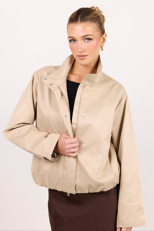 A woman modeling a beige lightweight jacket with a stand-up collar and snap-button front, worn over a black top and brown skirt against a plain white studio background.