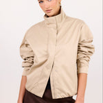 A woman modeling a beige wrap-style jacket with a high stand collar and gathered sleeves, paired with a dark brown skirt, against a plain white studio background.