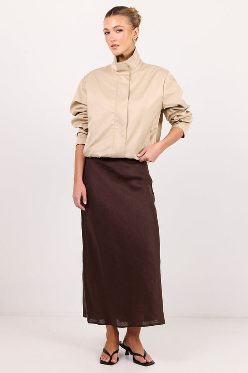 A woman modeling a beige mock-neck long-sleeve blouse and a dark brown midi skirt, standing against a plain white studio background.
