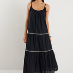 Woman wearing a plain black tiered maxi dress with strappy sleeves, white trim details, and back tie feature, styled with black sandals.