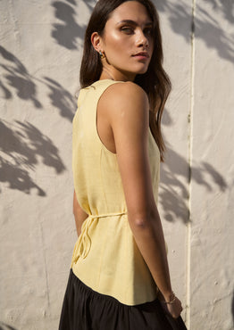 Woman wearing a sleeveless lemon yellow vest with a v-neckline and button-down front, standing against a sunlit wall with soft shadows cast from nearby foliage.