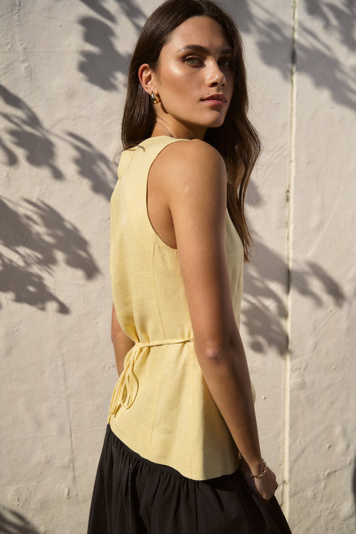 Woman wearing a sleeveless lemon yellow vest with a v-neckline and button-down front, standing against a sunlit wall with soft shadows cast from nearby foliage.
